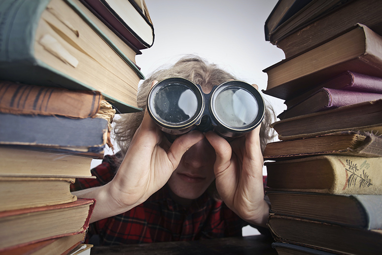 Man looking through binoculars behind a stack of books