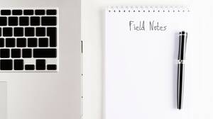 notepad next to a laptop with "Field Notes" written on the page and a pen on top of the page